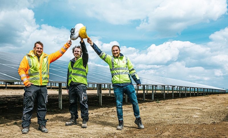 Karriere bei BELECTRIC: Drei Mitarbeitende stehen vor einer Solaranlage und halten ihre Sicherheitshelme in die Höhe. Sie lachen.
