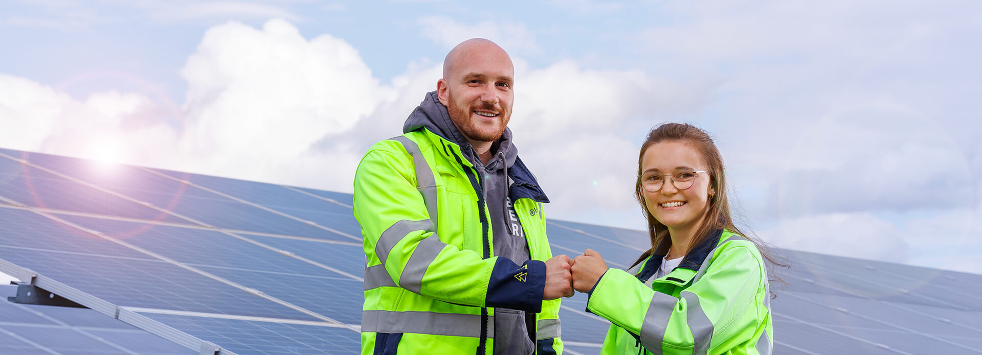 Two electricians working for solar energy company BELECTRIC fist-bump in front of a solar farm and smile