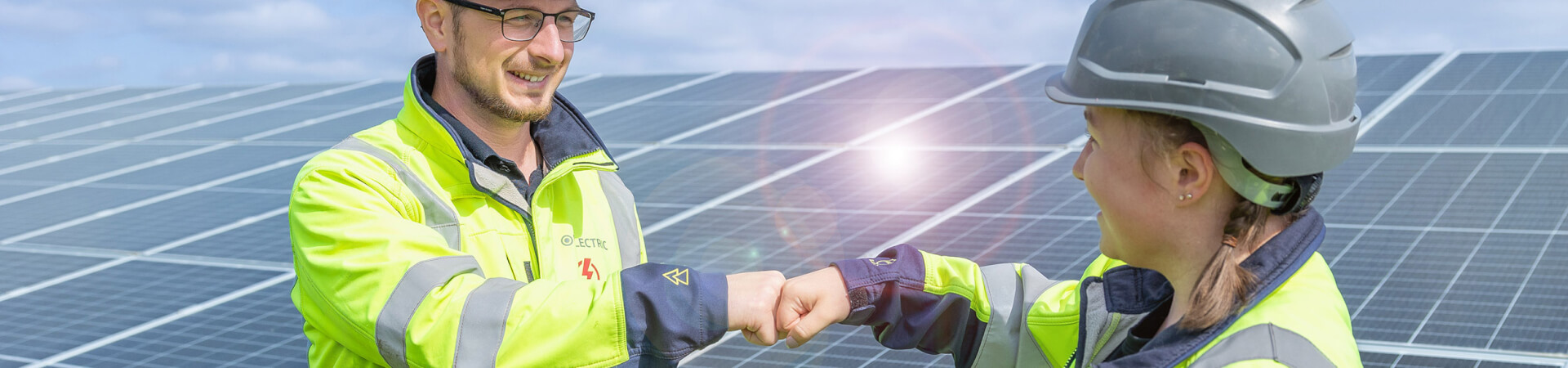 Two electricians working for a provider of solar systems fist-bump in front of a solar farm