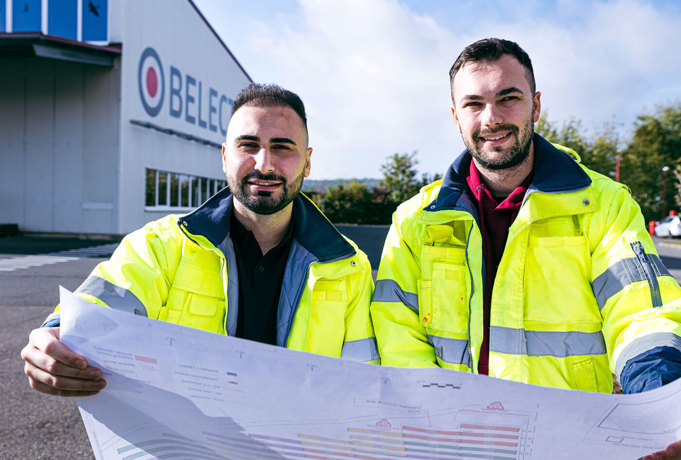 Two BELECTRIC employees in EPC wear warning jackets, hold a plan and smile into the camera