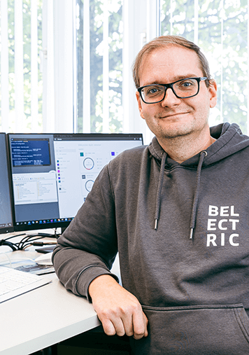 A BELECTRIC employee working in Technical Asset Management sits in front of his computer and smiles into the camera