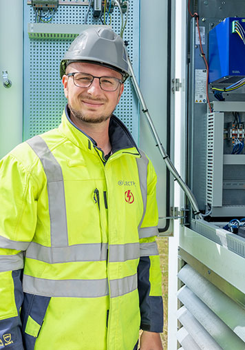 A BELECTRIC electrician working in Operations & Maintenance stands in front of an inverter and smiles into the camera