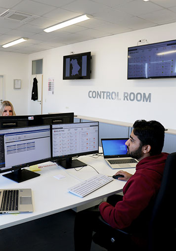 A BELECTRIC employee working in Operations & Maintenance sits in front of a computer. Behind him on the wall, there is a sign that says "Control Room".