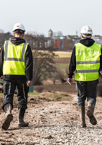 Two project developers working for BELECTRIC are walking across a field. They wear yellow warning vests.