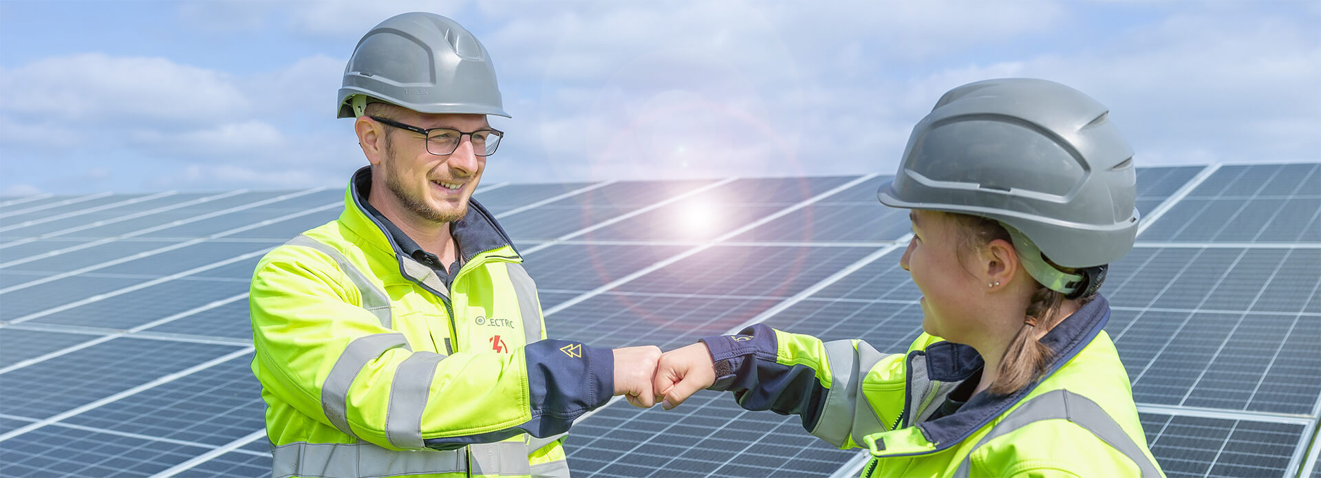 Two electricians working for solar energy company BELECTRIC fist-bump in front of a solar farm and smile