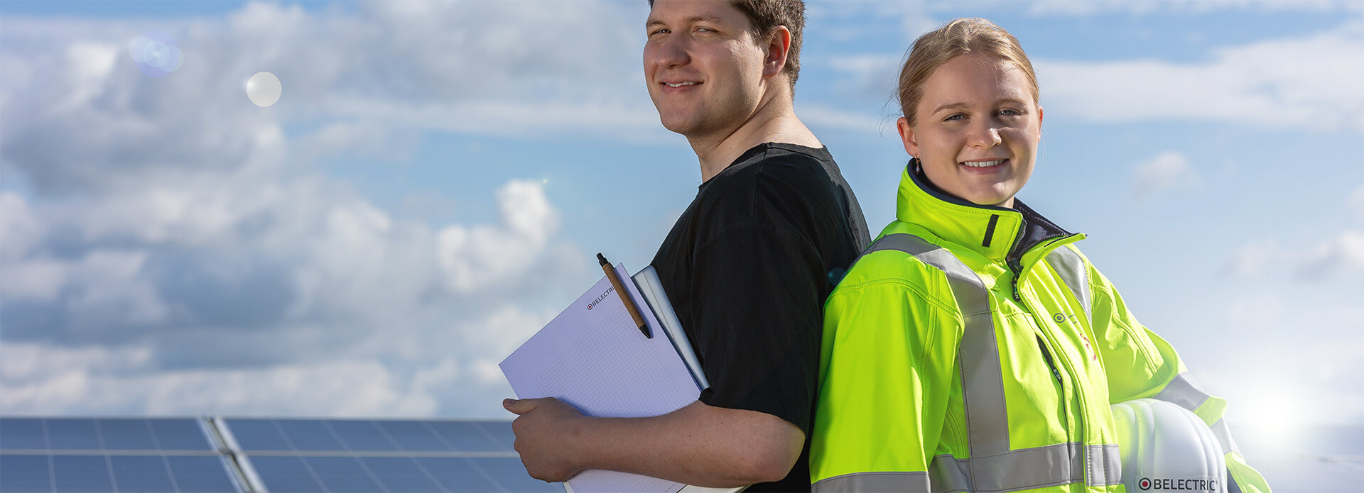 Two trainees working for solar energy company BELECTRIC stand in front of a solar farm. One of them is wearing a high-visability jacket and the other one is carrying a laptop, paper and pen.