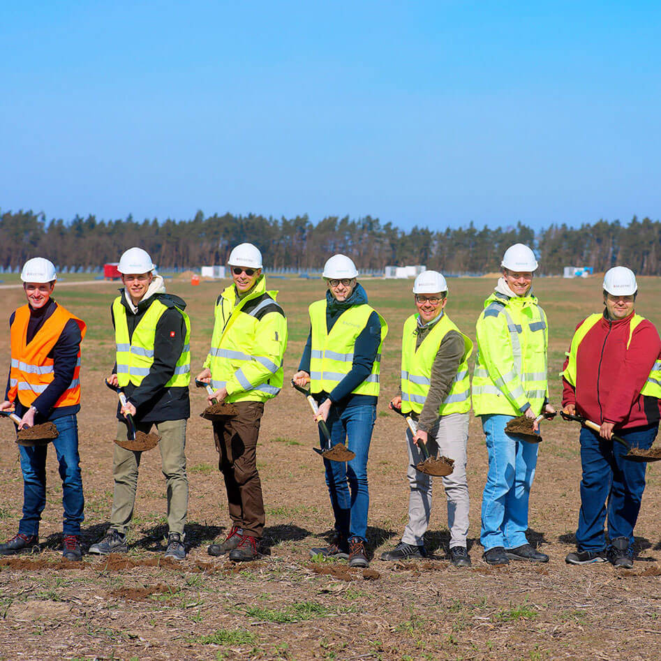 Seven people wearing high-visibility jackets stand on the Borrentin solar farm construction site and hold spades