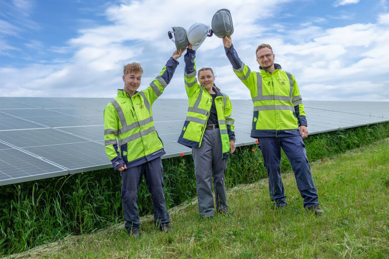 Three electricians wearing high-visibility jackets stand in front of a solar farm and hold up their safety helmets