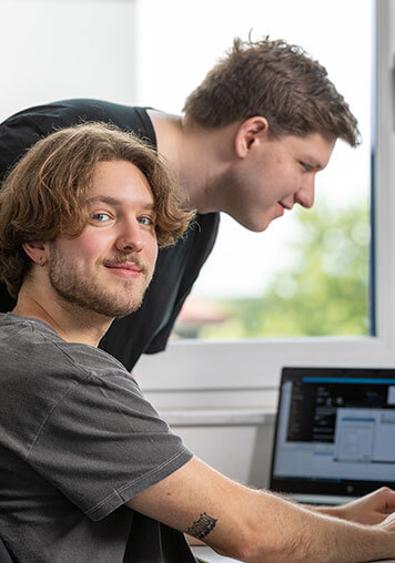 A BELECTRIC employee sits in front of a computer and smiles into the camera. Behind him, there is a second employee who looks at his monitor.