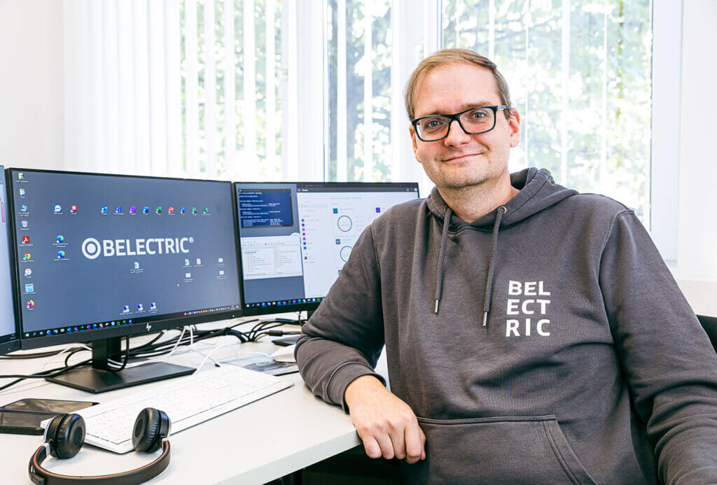 A male employee sits in front of his desk, where two monitors are placed. He smiles into the camera, wearing a branded hoodie that shows he pursues a career at BELECTRIC.