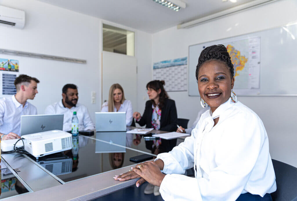 A female employee sits in front of a meeting table and smiles into the camera. In the background, several people are seen having a discussion in front of their laptops.