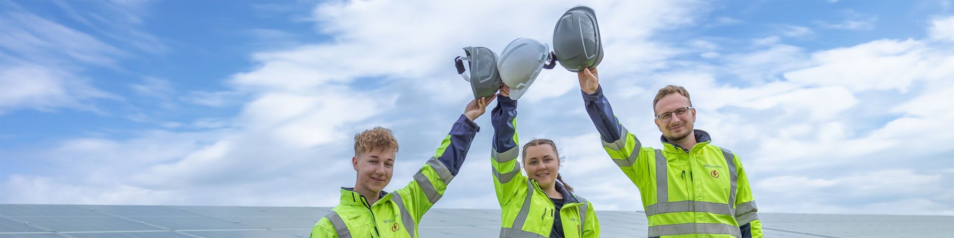 Three BELECTRIC employees stand in front of a solar farm and hold up their helmets.