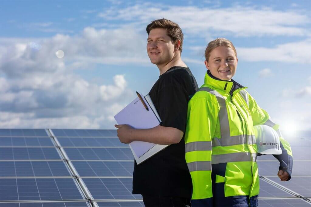 Two trainees stand in front of a solar farm. one of them is wearing a high-visibility jacket and holds a helmet. The second is holding a laptop, pen, and paper.