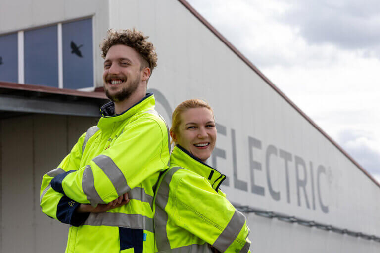 Two electrical engineering students stand in front of a building, wearing high-visibility jackets. They cross their arms and smile into the camera.