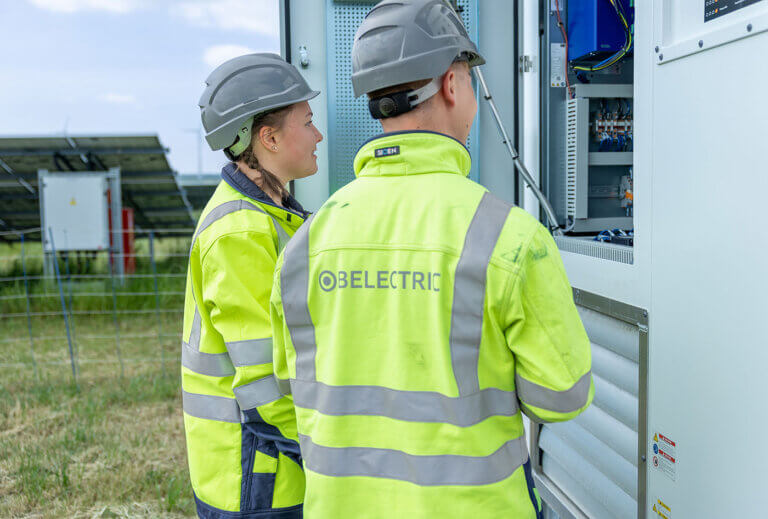 Two electronics technician trainees stand in front of an inverter on a solar farm.