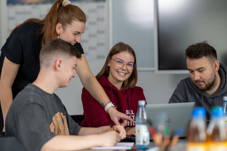 Several trainees sit around a meeting table, discussing, smiling and pointing towards a laptop in front of them.