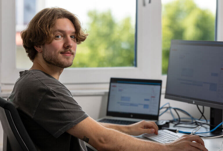 A BELECTRIC trainee training to become an IT specialist sits in front of a computer and smiles into the camera.