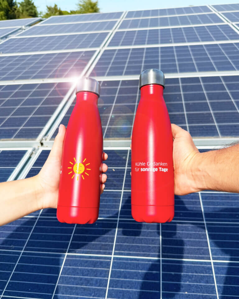 Zwei rote Thermosflaschen werden vor eine Solaranlage gehalten. Auf den Flaschen ist eine gelbe Sonne zu sehen sowie der Spruch "Kühle Gedanken für sonnige Tage"