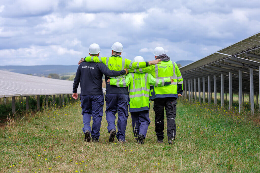 Four employees wearing high-visibility clothing with "BELECTRIC" branding walk across a solar farm, arm in arm