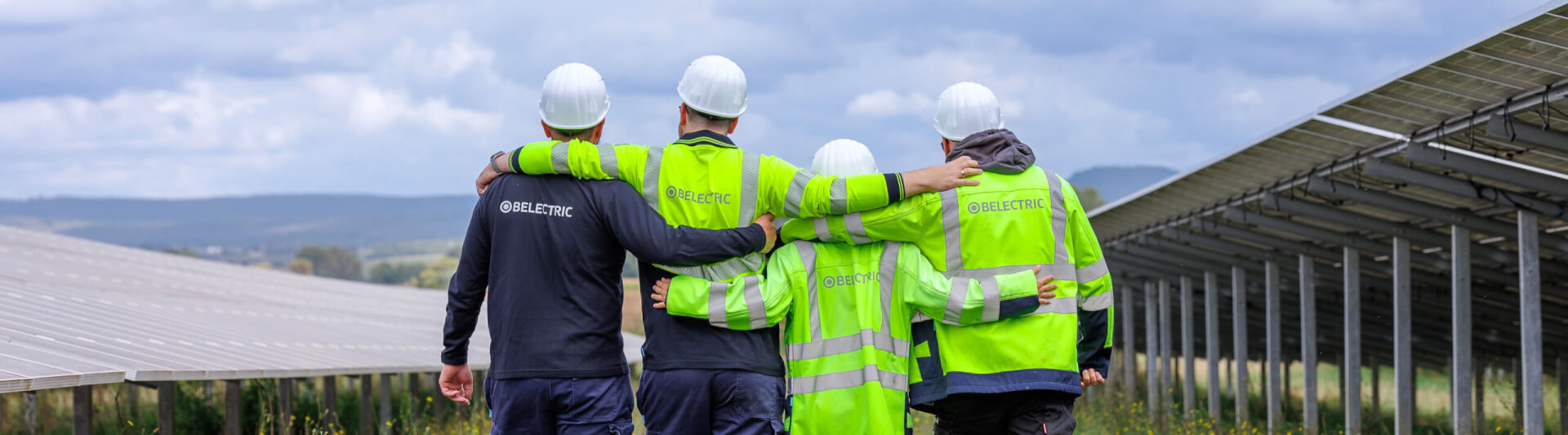 Vier BELECTRIC-Mitarbeitende in Warnjacken laufen Arm in Arm über einen Solarpark und feiern das 25. Jubiläum des Unternehmens