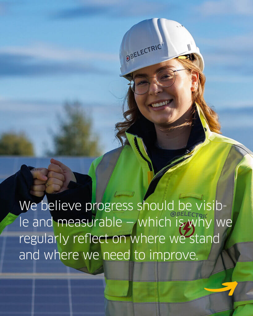 A female electrician wearing a high-vis jacket and helmet stands in front of a solar park. The text reads "We believe progress should be visible and measurable - which is why we regularly reflect on where we stand and where we need to improve".