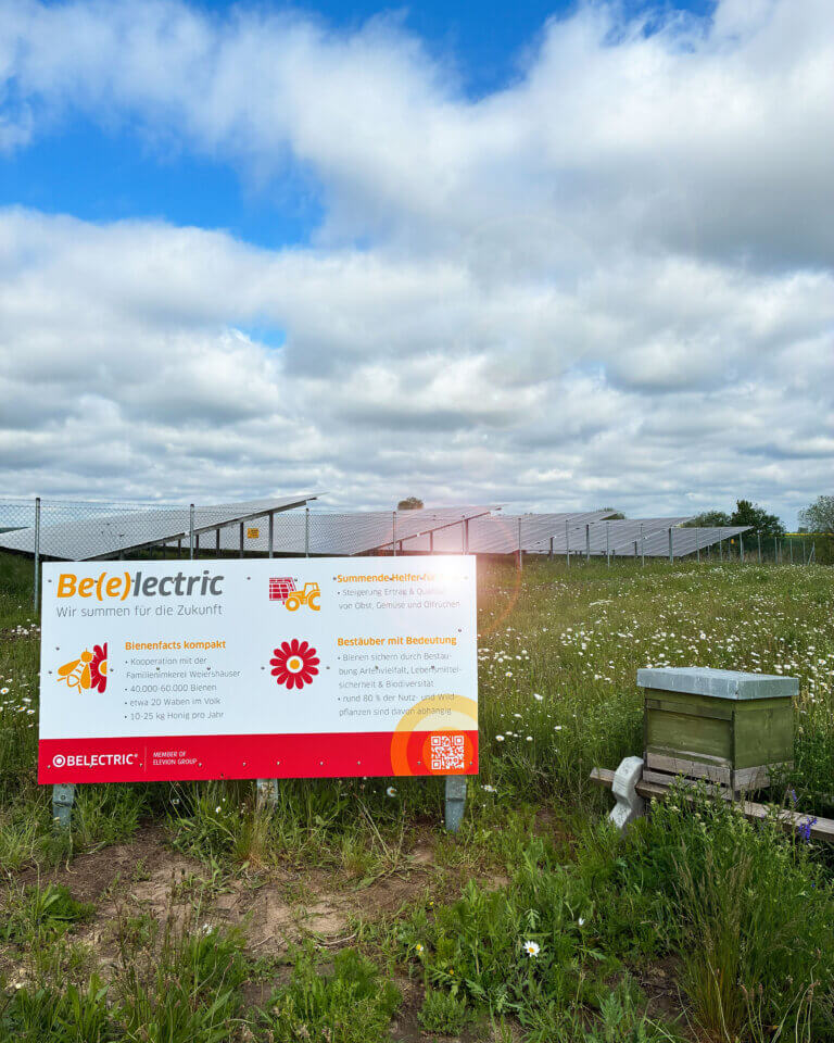 Ein Bienenstock und ein Schild vor einem Solarpark. Auf dem Schild steht: "Be(e)lectric: Wir summen für die Zukunft".