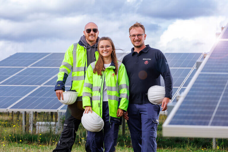 Three BELECTRIC employees stand on a solar farm, smiling into the camera