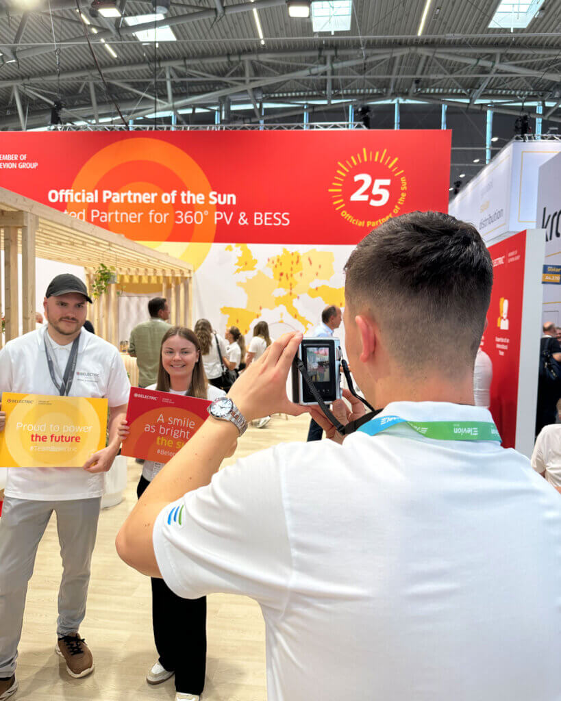 Zwei Gäste werden vor dem BELECTRIC-Stand auf der Intersolar mit einer Polaroidkamera fotografiert. Sie halten Schilder mit den Aufschriften „Proud to power the future“ und „A smile as bright as the sun“.