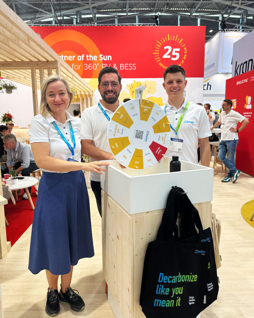 Drei Mitarbeitende von Elevion stehen hinter einem Glücksrad am BELECTRIC-Stand auf der Intersolar.